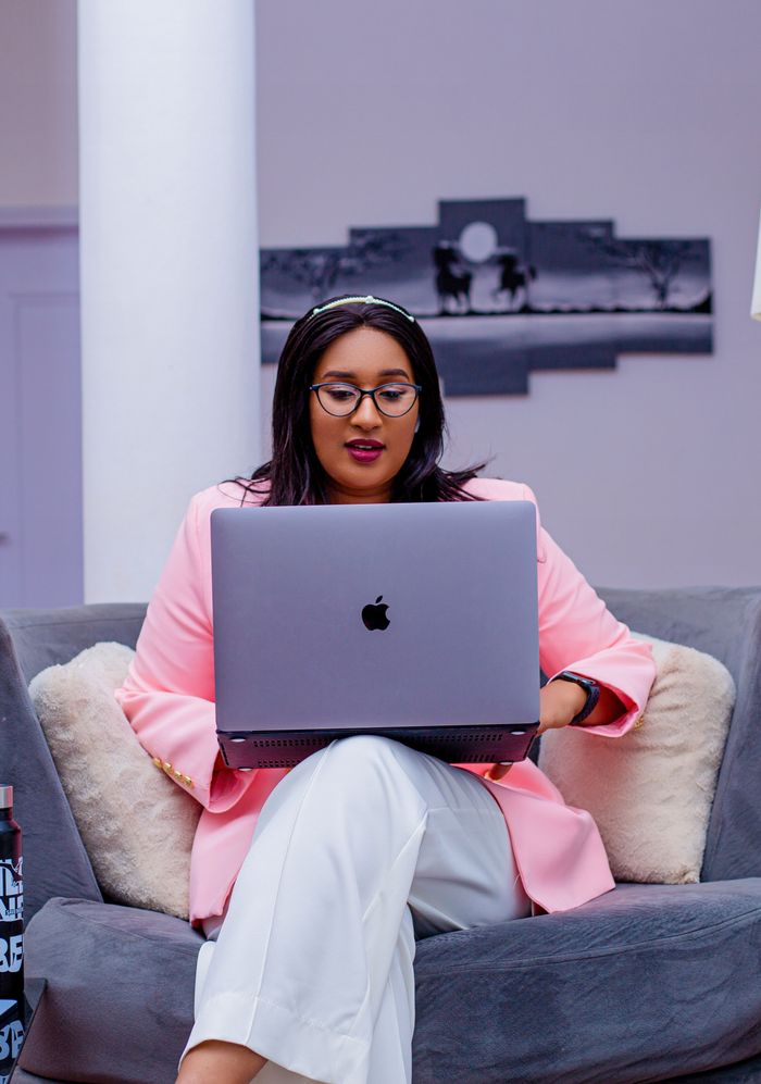 Focused woman in pink blazer works on laptop seated on a cozy gray couch indoors.